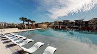 This outdoor pool area is designed to provide a full-scale recreational experience. The centerpiece is a generously sized swimming pool with clear, blue water, complemented by a separate spa or wading area nearby. The expansive pool deck is finished in smooth concrete and lined with an array of lounge chairs, including sleek, modern reclining sun loungers as well as traditional poolside seating, offering ample space for sunbathing and relaxation. Shade umbrellas are positioned throughout the deck, providing options for guests seeking relief from direct sunlight.

Mature palm trees and lush landscaping frame the perimeter, contributing a resort-like ambiance. The pool area benefits from abundant natural light and open sky views, enhancing the outdoor experience. A dedicated pool house structure serves as the entry point, featuring a stucco exterior, rustic dark-wood framed glass doors, and wall-mounted lighting. The pool house interior is compact and functional, offering direct visual access to the pool through its glass entryway.

The surrounding multi-story building complex provides an attractive architectural backdrop, and the overall layout is thoughtfully organized to maximize usability, comfort, and visual appeal for residents and guests throughout the day.