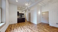 Located at 850 W St Clair Ave in Cleveland, Ohio, this single-floor loft-style unit showcases an industrial-chic aesthetic rooted in its converted building origins. The open-concept living and kitchen area is anchored by warm hardwood flooring, soaring ceilings with exposed white cylindrical ductwork, and a ceiling fan that enhances the airy, loft-like atmosphere. Painted brick walls add textural character throughout, reinforcing the space's urban industrial charm.

The kitchen is well-appointed with dark espresso cabinetry, granite countertops, stainless steel appliances including a refrigerator, range, microwave, and dishwasher, and a functional peninsula that serves as both a workspace and casual dining area. Recessed lighting provides consistent illumination across the open living space.

The bedroom features soft carpeting, a window with blinds that draws in natural light, painted brick walls, and an open closet with built-in shelving and hanging rods, offering practical storage. The bathroom is cleanly finished with hardwood-style flooring, a tub-shower combination, granite-topped dark vanity, and chrome fixtures. The overall ambiance is modern and urban, blending industrial character with contemporary finishes.