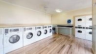 This shared laundry facility is thoughtfully designed to combine practicality with a welcoming aesthetic. The single-floor space is finished with light wood-look flooring that runs throughout, complementing the soft yellow upper walls and sage green beadboard wainscoting that wrap the room in a clean, cottage-inspired palette. Flush-mount ceiling fixtures provide bright, even lighting across the entire space.

Along one wall, a row of commercial-grade front-load washing machines is neatly lined up beneath a continuous countertop surface, offering convenient in-cycle monitoring and ample workspace. Additional dryer units are positioned nearby, rounding out a comprehensive laundry setup suited for high-capacity use. A central folding table with turned legs provides a dedicated surface for sorting and folding garments, while a built-in bench along the opposite wall offers comfortable seating while waiting.

A payment kiosk mounted on the wall provides a streamlined way to operate the machines. The facility is accessed through a standout decorative security door featuring an ornate sunburst metalwork design, allowing natural light to filter in while maintaining security. A window on the adjacent wall further brightens the space with natural light and a view of outdoor greenery. A fire extinguisher is visibly mounted near the entrance, underscoring the facility's attention to safety.