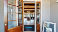 Located at Overall Creek Apartment Homes in Murfreesboro, Tennessee, this thoughtfully designed single-floor community clubhouse offers a generous layout across a well-appointed footprint. The entrance is framed by striking wood-and-glass French doors that open into a grand living area showcasing soaring ceilings with exposed wood beam accents, rich hardwood floors, and an eye-catching chandelier that anchors the space with warmth and sophistication. A linear fireplace set into a shiplap-clad accent wall, paired with a wall-mounted TV, creates an inviting focal point in the lounge area, which is bathed in natural light from multiple sets of French doors along the exterior wall. The kitchen features sage-green cabinetry, stainless steel appliances including a full-size refrigerator, microwave, and dishwasher, a mosaic tile backsplash, and a large island with quartz countertops and bar seating — all connected to the main living area through an arched passageway. Two distinct dining areas offer flexible meeting and gathering options, each illuminated by modern globe pendant chandeliers. Additional rooms include dedicated wellness spaces equipped with a tanning bed and a relaxation lounge chair with ambient lighting and acoustic panels, providing a spa-like retreat within the community.