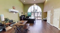 This leasing office makes a strong first impression with its dramatic architectural design, highlighted by a large, floor-to-ceiling arched window that floods the interior with abundant natural light and offers views of the surrounding landscaped grounds, including mature trees and desert vegetation. The soaring ceilings amplify the sense of openness throughout the main area, which is finished in warm yellow-green tones that lend an inviting, energetic atmosphere.

The main workspace features a well-appointed leasing desk equipped with computer workstations and ample storage, positioned to face the arched window for an airy, productive environment. A ceiling fan overhead adds to the comfort of the space. A rustic wood communal table with bench seating provides a casual area for meetings or informational sessions.

A welcoming reception nook near the entrance includes seating for visitors, decorative wall accents, and a chalkboard display for current leasing promotions. Wood-look flooring runs throughout, adding warmth and cohesion to the design.

Toward the rear of the office, additional private rooms are accessible through classic paneled doors set against warm gold-toned walls. A dedicated refreshment station with a coffee maker and red accent shelving offers a hospitable touch for guests. The overall ambiance is bright, professional, and approachable.