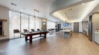 This contemporary amenity space is thoughtfully designed to offer a dynamic blend of recreation, relaxation, and productivity across a single open floor. The game and lounge area serves as the social centerpiece, featuring a full-size billiards table, plush gray sofas, and stylish black-and-gold accent chairs arranged around a flat-screen TV set into a dramatic dark-painted niche. Sunburst wall décor and gold-framed furnishings add a refined, upscale touch throughout. Floor-to-ceiling windows with roller shades flood the space with abundant natural light and offer views of the surrounding greenery, creating an airy, welcoming atmosphere. High-top bistro tables and bar-height seating provide casual gathering spots near the windows. A striking kitchen bar anchors one end of the space, showcasing an expansive marble-topped island with bar stools, pendant lighting, a patterned tile backsplash, glass shelving, stainless steel appliances, and a dedicated coffee station. A coffered ceiling with integrated cove lighting adds architectural distinction to this area. Wood-look flooring runs throughout, unifying the space with a warm, modern aesthetic. A separate, quietly styled workstation area with dark-wall accents completes this versatile and well-appointed amenity space.