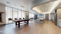 This contemporary amenity space is thoughtfully designed to offer a dynamic blend of recreation, relaxation, and productivity across a single open floor. The game and lounge area serves as the social centerpiece, featuring a full-size billiards table, plush gray sofas, and stylish black-and-gold accent chairs arranged around a flat-screen TV set into a dramatic dark-painted niche. Sunburst wall décor and gold-framed furnishings add a refined, upscale touch throughout. Floor-to-ceiling windows with roller shades flood the space with abundant natural light and offer views of the surrounding greenery, creating an airy, welcoming atmosphere. High-top bistro tables and bar-height seating provide casual gathering spots near the windows. A striking kitchen bar anchors one end of the space, showcasing an expansive marble-topped island with bar stools, pendant lighting, a patterned tile backsplash, glass shelving, stainless steel appliances, and a dedicated coffee station. A coffered ceiling with integrated cove lighting adds architectural distinction to this area. Wood-look flooring runs throughout, unifying the space with a warm, modern aesthetic. A separate, quietly styled workstation area with dark-wall accents completes this versatile and well-appointed amenity space.