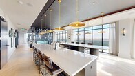 This expansive, single-floor amenity space is designed with a refined contemporary aesthetic, seamlessly blending social, dining, and work-focused environments across a generously proportioned layout. The interior is anchored by a striking open kitchen and bar area featuring an elongated white quartz island illuminated by a series of cascading gold pendant lights suspended from a dark ceiling plane. Light chevron-patterned hardwood floors run throughout, providing a warm contrast to the deep navy and charcoal architectural accents defining the space.

Floor-to-ceiling curtain wall windows dominate multiple elevations, flooding the interior with natural light and framing panoramic city skyline views. A dedicated lounge zone offers a curated arrangement of contemporary seating, including swivel chairs with gold-toned bases, modular sofas, and built-in banquette booths with dark upholstery and slatted wood accent panels. A large built-in media wall with integrated shelving serves as a dramatic focal point.

Private workspaces include glass-enclosed offices with textured feature walls and individual workstations, as well as semi-private booth-style nooks along a wide corridor. A dedicated conference room with direct outdoor terrace access and skyline views provides a focused meeting environment. Throughout, curated artwork and thoughtful material selections contribute to a cohesive, upscale ambiance.
