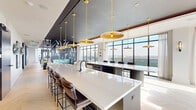 This expansive, single-floor amenity space is designed with a refined contemporary aesthetic, seamlessly blending social, dining, and work-focused environments across a generously proportioned layout. The interior is anchored by a striking open kitchen and bar area featuring an elongated white quartz island illuminated by a series of cascading gold pendant lights suspended from a dark ceiling plane. Light chevron-patterned hardwood floors run throughout, providing a warm contrast to the deep navy and charcoal architectural accents defining the space.

Floor-to-ceiling curtain wall windows dominate multiple elevations, flooding the interior with natural light and framing panoramic city skyline views. A dedicated lounge zone offers a curated arrangement of contemporary seating, including swivel chairs with gold-toned bases, modular sofas, and built-in banquette booths with dark upholstery and slatted wood accent panels. A large built-in media wall with integrated shelving serves as a dramatic focal point.

Private workspaces include glass-enclosed offices with textured feature walls and individual workstations, as well as semi-private booth-style nooks along a wide corridor. A dedicated conference room with direct outdoor terrace access and skyline views provides a focused meeting environment. Throughout, curated artwork and thoughtful material selections contribute to a cohesive, upscale ambiance.