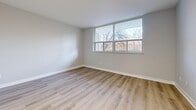 This efficiently designed single-floor unit comprises four well-defined spaces: a living room, kitchen, hallway, and bathroom, all unified by a consistent aesthetic of clean lines and modern finishes. Light-toned hardwood-style flooring runs throughout, lending warmth and visual continuity to the entire unit.

The living room is the most spacious area, featuring soft gray walls, crisp white baseboards, and a large multi-pane window that invites an abundance of natural light and offers views of mature trees outdoors. Multiple electrical outlets are conveniently positioned along the walls.

The kitchen is thoughtfully appointed with white shaker-style cabinetry — both upper and lower — complemented by granite-patterned countertops in shades of white and gray. A stainless steel sink is integrated into the counter, and the layout makes efficient use of the available space.

The hallway connects the rooms with ease, featuring a mirrored sliding-door closet that adds depth and practicality. Additional doors along the hall provide access to the bathroom and other areas of the unit.

The bathroom is finished with white tile surrounding a full bathtub and shower combination, fitted with polished chrome fixtures. The clean, minimalist design maintains the unit's overall bright and neutral aesthetic. Together, these spaces create a functional and cohesive living environment.