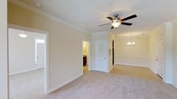 Located at 485 Traebert Pl in Raleigh, NC, this single-story space presents a well-appointed interior with a cohesive, classic design aesthetic. The main living area is finished with plush wall-to-wall carpeting in a neutral tone, creating a warm and inviting atmosphere. Crown molding runs along the ceiling perimeter, adding a refined architectural detail that elevates the overall character of the space.

Ceiling fixtures are thoughtfully selected, including a multi-blade ceiling fan with integrated lighting in the primary room, promoting both comfort and functionality. An elegant chandelier is visible in the adjoining area, lending a more formal ambiance. The rooms are connected through clean white-framed doorways, offering an open and accessible flow throughout the layout.

Walls are painted in soft, warm beige tones that complement the light-colored trim and white baseboards, contributing to a bright and airy feel. A louvered closet door adds a traditional design touch, while a glimpse into an adjoining room reveals dark wood cabinetry, suggesting a well-finished secondary space. Natural light filters through windows, further enhancing the welcoming character of the interior.