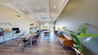 Located at 509 Stone Dr in Manhattan, Kansas, this single-story community clubhouse offers a well-appointed gathering space designed to serve residents across multiple functional areas. The main lounge is the heart of the facility, boasting soaring ceilings with elegant coffered detailing, rich hardwood-style flooring, and an abundance of natural light streaming through a dramatic arched window. Multiple seating and dining arrangements provide versatile spaces for socializing or casual meetings, complemented by warm, neutral tones and tasteful furnishings throughout. A dedicated business area features a wall-mounted countertop workstation with computers, ideal for residents needing a focused work environment. The fully equipped kitchen includes ample white cabinetry, dark granite-style countertops, a stainless steel sink, and a full-size refrigerator, making it well-suited for community events and gatherings. The fitness center is thoughtfully divided into two areas — one housing cardio equipment including treadmills, an elliptical, and cycling bikes alongside a floor-to-ceiling mirror wall, and another open floor space stocked with medicine balls, resistance equipment, and yoga mats. Together, these amenities create a comprehensive, inviting community hub.