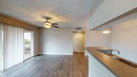 This single-story residence features a well-designed open-concept layout connecting the living area and kitchen. The living room is finished with durable wood-style flooring, a textured ceiling, and a ceiling fan with integrated lighting. A sliding glass door with vertical blinds allows natural light to fill the space and provides outdoor access. The kitchen is configured in a galley style, featuring white cabinetry, dark granite-style countertops, a stainless steel refrigerator, an electric range, a dishwasher, and a double-basin sink beneath a window. A peninsula with a dark countertop separates the kitchen from the living area, with pendant lighting above. The front entry, framed by a door with sidelight windows, opens directly into the main living space. The bathroom includes a tub-shower combination with white tile surround, a white vanity with a stone-style countertop, a large mirror, and wood-style flooring. The bedroom features plush carpet, a ceiling fan, a textured ceiling, and a large window offering natural light. Throughout the unit, neutral tones and consistent finishes create a cohesive and functional living environment.