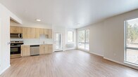 Located in Bend, Oregon, this two-story residence offers a clean, contemporary design across two well-proportioned floors. The main level features an open-concept kitchen and living area bathed in natural light from large windows overlooking lush greenery. The kitchen is equipped with stainless steel appliances, including an electric range, microwave, and dishwasher, complemented by light wood cabinetry, white quartz countertops, and a sink. Light-toned luxury vinyl plank flooring flows throughout the main level, enhancing the bright, airy atmosphere. A private exterior door provides direct outdoor access from the living area. The upper floor houses two bedrooms, including a generously sized primary suite, both carpeted for comfort and featuring windows with natural views. The primary bedroom connects to an accessible bathroom with wood-toned vanity cabinetry, white quartz countertops, tile shower, and grab bars. The residence includes five bathrooms in total, each finished with consistent, modern fixtures. A staircase connects the two levels, and a dedicated hallway and closet add to the home's practical layout. The overall aesthetic is bright and minimalist, with neutral tones, recessed lighting, and clean lines throughout.
