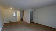 This multi-level residence is thoughtfully arranged across three floors, offering a well-organized layout suited for comfortable everyday living. The main living area features wall-to-wall carpeting in a warm neutral tone, providing a cozy and inviting atmosphere throughout. The textured ceiling adds a classic architectural detail, while the clean white walls create a bright, open feel that allows for versatile interior styling.

A central staircase connects the floors, framed by a dividing wall that contributes to the functional flow of the space. A secondary staircase is also visible, suggesting convenient access between multiple levels. Near the entryway, a wall-mounted mirror adds a practical touch and helps reflect light throughout the space.

The entryway corridor, highlighted by a warm-toned door, provides a welcoming transition into the main living areas. The layout efficiently separates living and transitional zones, maximizing the usability of each floor. Overall, the space presents a straightforward, practical design with neutral finishes throughout, making it an adaptable home ready for personalization. The three-floor configuration provides ample room for distinct living, sleeping, and utility areas across its levels.