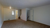 This multi-level residence is thoughtfully arranged across three floors, offering a well-organized layout suited for comfortable everyday living. The main living area features wall-to-wall carpeting in a warm neutral tone, providing a cozy and inviting atmosphere throughout. The textured ceiling adds a classic architectural detail, while the clean white walls create a bright, open feel that allows for versatile interior styling.

A central staircase connects the floors, framed by a dividing wall that contributes to the functional flow of the space. A secondary staircase is also visible, suggesting convenient access between multiple levels. Near the entryway, a wall-mounted mirror adds a practical touch and helps reflect light throughout the space.

The entryway corridor, highlighted by a warm-toned door, provides a welcoming transition into the main living areas. The layout efficiently separates living and transitional zones, maximizing the usability of each floor. Overall, the space presents a straightforward, practical design with neutral finishes throughout, making it an adaptable home ready for personalization. The three-floor configuration provides ample room for distinct living, sleeping, and utility areas across its levels.