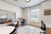 Located at 400 Southwest Pkwy in College Station, Texas, this single-room business center is thoughtfully designed to support both focused individual work and small group collaboration. The space features a built-in workstation along one wall, equipped with a desktop computer and a light-toned laminate desk surface, accompanied by an ergonomic task chair. A multifunction printer station sits conveniently nearby, making the area fully functional for everyday office needs. A round conference table with stylish black and gold chairs occupies the opposite side of the room, providing a comfortable setting for small meetings or group discussions. The walls are finished in a clean, neutral palette with crisp crown molding details, contributing to a polished and professional atmosphere. Large windows fitted with horizontal blinds allow ample natural light to filter in while offering views of the surrounding greenery. A large interior window opens onto an adjacent lounge or common area, enhancing the sense of openness and connectivity. Light-toned patterned carpet covers the floor throughout, adding warmth and texture to the space. Framed abstract artwork adorns the walls, lending a contemporary aesthetic. The overall ambiance is bright, modern, and functional — well-suited for productivity in a community-oriented setting.