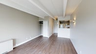 This single-floor unit presents a thoughtfully designed open-concept layout that maximizes both functionality and visual appeal. The main living area is defined by distinctive exposed ceiling beams that add architectural character to the space, complemented by neutral taupe-toned walls and warm hardwood-style flooring that runs throughout. Natural light enhances the bright, airy atmosphere, contributing to a clean and contemporary ambiance.

A built-in closet with sliding white doors provides convenient storage within the living area, while an entry corridor leads to additional rooms beyond. The pass-through kitchen is positioned in the corner of the unit, featuring overhead cabinetry, stainless steel appliances including a refrigerator, and a raised countertop bar that opens toward the main living space — ideal for casual dining or entertaining. Wall-mounted lighting fixtures flank the kitchen opening, adding both function and a modern aesthetic touch.

The overall design emphasizes simplicity and practicality, with white trim, clean lines, and a cohesive neutral palette tying each element together. The open flow between the kitchen and living areas creates a sense of spaciousness well-suited to a variety of living arrangements.