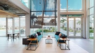 Located at 100 E Tyler St. in Tampa, FL, this contemporary single-floor amenity space combines striking architectural design with a diverse range of social and recreational functions. The entrance lounge immediately impresses with soaring double-height floor-to-ceiling glass walls that flood the space with natural light and frame sweeping views of the surrounding palm-lined Tampa streetscape. Teal sofas, warm wood-accented side tables, and light tile flooring create a welcoming, resort-inspired atmosphere. Adjacent to the lounge, a vibrant media and seating area offers a wall-mounted flat-screen TV, bold red upholstered chairs, and colorful artwork, fostering an inviting environment for casual gatherings. A sleek, well-appointed kitchen area features expansive white countertops, a wood slat ceiling detail, industrial-style pendant lighting, and terrazzo flooring, providing both functionality and visual warmth. The space transitions seamlessly into a lively game room complete with foosball, a pool table, multiple flat-screen displays, and eclectic hexagonal patterned carpet tiles in red and gray. Throughout, frosted glass partitions and open sightlines maintain a sense of connectivity while defining distinct zones, making this a versatile and energetic communal destination in the heart of Tampa.
