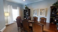 This single-floor space is designed with a refined and welcoming aesthetic, highlighted by a beautifully appointed dining area. The dining room centers around a substantial dark wood table surrounded by high-backed leather chairs with nailhead trim detailing, offering both comfort and classic style. The walls are finished in a soft, muted blue tone that lends a calm and sophisticated ambiance to the room. Natural light filters in through a large window dressed with sheer white curtains, brightening the space and adding an airy quality. A dark wood bookcase with ornate detailing serves as a prominent decorative feature, topped with lush greenery and displaying carefully curated decorative objects. The walls are adorned with framed botanical and floral artwork in complementary blue and neutral tones, enhancing the cohesive color palette throughout. Crown molding adds an additional layer of architectural refinement, framing the ceiling and contributing to the overall sense of elegance. Neutral carpeting grounds the room and adds warmth underfoot. Together, these elements create a space that is both functionally suited for gathering and visually polished, making it ideal for formal dining or meetings in a comfortable, well-designed setting.
