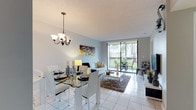 This well-appointed single-floor residence offers a thoughtfully designed layout that balances comfort and functionality. The open-plan living and dining area serves as the heart of the home, featuring light-toned walls, gleaming white tile flooring, and an elegant chandelier above the glass-top dining table with modern white chairs. The living area flows seamlessly toward a full-width sliding glass door that opens onto a private balcony, flooding the interior with natural light and offering pleasant views of surrounding greenery and palm trees.

The kitchen is clean and efficient, outfitted with white cabinetry, granite countertops, tile flooring, and a full suite of white appliances including a refrigerator, dishwasher, and range. The bedrooms are carpeted and finished in neutral tones, providing a calm retreat. The primary bedroom features direct balcony access through a sliding glass door, a closet, and an en-suite bath. A secondary bedroom offers comfortable space with large windows. The bathrooms are tiled throughout, with granite vanity tops, a tub-shower combination, and warm accent walls. An additional flexible room with carpeting and a window provides space adaptable for varied needs.