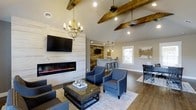 This inviting single-floor space is thoughtfully designed to blend rustic warmth with contemporary style. The centerpiece of the main lounge area is a sleek, wall-mounted electric fireplace set against a striking white shiplap accent wall, creating a cozy yet modern focal point. Above, vaulted ceilings showcase exposed wooden beam details that add architectural character and a sense of warmth throughout the space. A rustic chandelier with candle-style lighting hangs gracefully overhead, complementing the natural wood tones found in the beams and hardwood flooring. The lounge is furnished with navy blue upholstered armchairs accented with nailhead trim, arranged around a glass-top coffee table atop a neutral area rug, creating a comfortable and well-appointed seating arrangement. Adjacent to the lounge, a bar area features stone accent walls, pendant lighting, and bar stool seating, making it ideal for casual gatherings. Recessed lighting and a ceiling fan ensure both functional illumination and climate comfort. Natural light filters in through windows, brightening the space and highlighting its carefully chosen finishes. Overall, the space exudes a welcoming, upscale ambiance suited for socializing and relaxation.