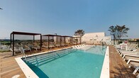 This rooftop pool deck is a thoughtfully designed outdoor amenity space positioned high above the city, offering striking panoramic views of the Washington, D.C. skyline, including a clear sightline to the U.S. Capitol dome. The centerpiece is a generously sized rectangular swimming pool with clear turquoise water, accessible via stainless steel entry rails and steps. The surrounding deck is finished in warm-toned hardwood planking, creating an inviting and cohesive aesthetic throughout the space.

A row of modern pergola structures, framed in deep brown metal with horizontal slatted louvers, lines one side of the deck and provides semi-shaded retreats equipped with cushioned lounge chairs. Additional open-air loungers with neutral mesh seating are arranged in clusters around the perimeter, ideal for sunbathing while taking in the expansive urban skyline. Accent trees punctuate the space, adding a natural, softening element to the contemporary design.

A standout feature is a large-scale, vividly colored mural adorning one wall of the rooftop, depicting bold botanical and wildlife imagery in bright reds, blues, and greens, adding an artistic focal point to the space. The combination of open sky, city views, curated greenery, and artistic details gives this rooftop deck a dynamic and resort-like atmosphere.