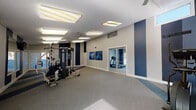 This fitness center is designed to support a comprehensive workout experience across its single-floor layout. The space is divided into distinct functional zones, seamlessly connected through glass-paneled double doors that maintain an open, airy feel while defining separate areas for different types of exercise.

The main workout area features a multi-station cable and strength machine as its centerpiece, offering a wide range of resistance training options. A stationary exercise bike and treadmill round out the cardio equipment, providing members with versatile options for aerobic training. Motivational fitness artwork adorns the walls, reinforcing the active atmosphere throughout the space.

The interior design is defined by a striking navy blue and white color scheme, with bold vertical navy stripes accenting the light gray walls. Striped carpet in coordinating shades of blue and gray flows across the floors, adding visual interest and delineating zones within the open layout. Ceiling fans and bright flush-mounted lighting panels ensure consistent illumination and comfort during workouts.

Large sliding glass doors and windows allow natural light to fill portions of the space, offering views to the outdoors. HVAC vents are integrated into the ceiling for climate control, and the textured ceilings add a classic finishing touch. The overall ambiance is clean, energetic, and purposefully designed to motivate and accommodate a variety of fitness activities.