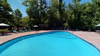 Located at 1105 NC-54 Hwy Bypass in Chapel Hill, NC, this single-level outdoor community amenity space offers a well-rounded recreational experience set amid lush, mature trees and manicured green surroundings. At the heart of the space is a generously sized, freeform swimming pool with vibrant blue water, flanked by lounge chairs and a shaded pergola structure — ideal for relaxing on warm Carolina days. Adjacent to the pool, a covered pavilion serves as a central gathering hub, equipped with a substantial stone-built outdoor grill, multiple dining tables with seating, and string lighting overhead, creating an inviting atmosphere for outdoor entertaining. The stone-paved patio beneath the pavilion extends the usable area and connects seamlessly to the surrounding landscape. A separate covered lounge area offers comfortable wicker seating with views of an open, expansive lawn, where mature trees provide a natural backdrop and a sense of privacy. The open green space beyond includes what appears to be a recreational court area, adding to the variety of outdoor activities available. The brick residential buildings visible in the background lend a classic, established character to the community. Overall, this amenity space is thoughtfully designed to encourage outdoor living, socializing, and recreation in a well-maintained, nature-rich setting.