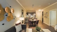 This single-floor residence offers a thoughtfully designed open-concept layout where the living, dining, and kitchen areas flow together with ease and elegance. The living area is anchored by a comfortable sofa and complemented by warm ambient lighting, creating an inviting atmosphere ideal for relaxing or entertaining. Crown molding adds a refined architectural touch throughout the space, enhancing the overall sense of sophistication.

The dining area features a round table paired with rich, plum-toned upholstered chairs, positioned beneath stylish pendant lighting that adds a warm, intimate glow. Hardwood flooring runs seamlessly across the living and dining areas, grounding the space with a sense of warmth and continuity.

The kitchen is a standout feature, boasting dark cabinetry, stainless steel appliances, a microwave, and a breakfast bar with barstool seating — a perfect space for casual meals or meal preparation. Track lighting along the kitchen ceiling provides bright, functional illumination. The neutral wall tones throughout complement the warm lighting scheme, creating a cohesive and inviting ambiance. A hallway leading from the main living area suggests additional private spaces beyond, completing this well-appointed and stylishly furnished residence.