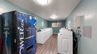 This shared laundry facility is a single-room, single-floor space designed to efficiently serve a multi-unit building. The room is organized with two parallel rows of commercial-grade top-load washers and dryers lining both side walls, creating a clear central walkway that maximizes accessibility and traffic flow throughout the space.

The interior features wood-look vinyl plank flooring that runs the length of the room, providing a clean and durable surface suited for high-traffic use. The walls are painted in a muted teal tone, giving the utilitarian space a degree of visual character. Overhead, flush-mount ceiling fixtures provide consistent artificial lighting throughout.

A beverage vending machine — stocked with Powerade and Dasani products — is positioned near the entrance, offering added convenience for users during longer laundry sessions. A small folding table is available along one wall, providing a surface for sorting or folding laundry. Informational signage, including laundry room rules and machine operating instructions, is posted at multiple points.

A single window on the rear wall allows a modest amount of natural light into the space. The room is accessed through a standard door that connects to a shared building corridor, with no-smoking signage posted at the entry.