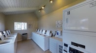 Located at 11355 Zapata in San Diego, this shared laundry facility offers a clean, functional, and well-maintained space designed to serve residents with ease and convenience. The single-floor layout features a charming vaulted ceiling with a white wood-paneled finish, lending the room a sense of openness and height that extends beyond its footprint. Track lighting mounted along the ceiling peak provides focused, ample illumination throughout the space. A row of white top-loading washing machines lines one wall, each equipped with individual card or coin-operated payment systems for user convenience. Stacked dryer units are positioned along the adjacent wall, maximizing the use of vertical space and accommodating multiple users simultaneously. The walls are finished in a neutral, muted tone that contributes to the room's clean and orderly atmosphere. A small window on one wall allows for a touch of natural light, while posted service information ensures residents have easy access to support resources. The carpeted flooring and a conveniently placed waste bin complete the tidy, practical environment. Overall, this laundry facility is thoughtfully organized to offer a straightforward, comfortable experience for everyday use.