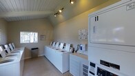 Located at 11355 Zapata in San Diego, this shared laundry facility offers a clean, functional, and well-maintained space designed to serve residents with ease and convenience. The single-floor layout features a charming vaulted ceiling with a white wood-paneled finish, lending the room a sense of openness and height that extends beyond its footprint. Track lighting mounted along the ceiling peak provides focused, ample illumination throughout the space. A row of white top-loading washing machines lines one wall, each equipped with individual card or coin-operated payment systems for user convenience. Stacked dryer units are positioned along the adjacent wall, maximizing the use of vertical space and accommodating multiple users simultaneously. The walls are finished in a neutral, muted tone that contributes to the room's clean and orderly atmosphere. A small window on one wall allows for a touch of natural light, while posted service information ensures residents have easy access to support resources. The carpeted flooring and a conveniently placed waste bin complete the tidy, practical environment. Overall, this laundry facility is thoughtfully organized to offer a straightforward, comfortable experience for everyday use.