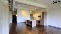 Located in Dallas, TX, this single-story space showcases an open-concept layout that seamlessly blends modern finishes with industrial design elements. The interior is anchored by rich, dark hardwood flooring that extends throughout, adding warmth and character to the space.

The kitchen is a standout feature, outfitted with contemporary wood-toned cabinetry in a two-tone configuration — combining lighter upper cabinets with darker lower and tall cabinet panels. A central island with a light countertop provides additional workspace and serves as a natural dividing point between the kitchen and the open living area. Stainless steel appliances, including a refrigerator, microwave, and range, complete the functional kitchen setup.

Overhead, exposed structural beams run across the ceiling, reinforcing the industrial aesthetic. Track lighting is installed along the ceiling, offering flexible, directed illumination throughout the open space. The expansive, unobstructed layout allows for versatile furniture arrangement and creates a spacious, airy feel. A corridor visible along the left side suggests additional areas beyond the main open space, contributing to the overall functionality of the unit.