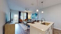 This thoughtfully designed single-floor residence offers a seamless blend of modern aesthetics and functional living. The open-concept layout connects the entryway, kitchen, dining, and living areas, creating a bright and cohesive flow throughout the main living space. Warm wood-look flooring runs throughout the common areas, complementing the soft, neutral color palette on the walls.

The kitchen is well-equipped with stainless steel appliances, sleek white cabinetry, light stone countertops, and a central island with a built-in sink — ideal for both everyday cooking and entertaining. Pendant lighting above the island adds a refined touch. The living area is generously proportioned, bathed in natural light from large windows dressed with navy curtains, and features a ceiling fan overhead.

The bedroom is a calm retreat, carpeted for comfort and illuminated by multiple windows that frame serene outdoor views. Ample natural light fills the room throughout the day. A hallway leads to the bathroom, which features wood-look flooring, a vanity with mirror, and clean, modern fixtures. The overall ambiance of the space is contemporary and welcoming, with consistent attention to detail evident in every room.
