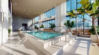This sophisticated amenity level is defined by its airy, light-filled architecture and panoramic urban views. Floor-to-ceiling windows and expansive glass curtain walls run along the perimeter, flooding the interiors with natural light and framing striking city skyline vistas throughout the day.

At the heart of the space is a generously sized indoor lap pool, elevated on a tiled platform and accessed via stainless steel handrails. The pool area features soaring ceilings with globe pendant lighting, sheer white drapery softening the glass walls, and lush potted greenery adding warmth to the refined palette of white tile and neutral stone flooring.

Adjacent to the pool, a sun deck lounge area is furnished with sleek reclining chaise lounges and low tables, bathed in natural sunlight streaming through the floor-to-ceiling windows. The deck connects seamlessly to the pool via steel-framed glass doors, creating a cohesive indoor-outdoor flow.

A separate lounge area showcases textured watercolor-inspired wallcovering, woven accent chairs, warm wood cabinetry, and curated artwork, offering a more intimate retreat. Shower facilities with grey subway tile complete the amenity suite. Throughout, lush tropical plants and carefully considered lighting elevate the overall ambiance.