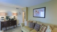 This single-floor space features a welcoming living area finished in soft, sage-green tones that create a calm and relaxed atmosphere. The living room is anchored by a plush, olive-toned sofa adorned with a collection of patterned throw pillows in earthy hues of brown, rust, and cream, adding texture and visual interest. A textured popcorn ceiling and light-toned carpet contribute to the room's cozy character. A framed floral artwork with bold poppy motifs serves as a focal point on the main wall, complementing the warm color palette throughout. A classic side table with a table lamp provides soft ambient lighting, while a small decorative console with dark wood tones adds a traditional accent. HVAC vents are integrated into both the walls and baseboards, ensuring year-round comfort. A hallway visible from the living area leads to additional rooms, with framed wall art lining the corridor, hinting at a thoughtfully decorated interior. The overall ambiance is warm, understated, and functional, making the space well-suited for comfortable everyday living.