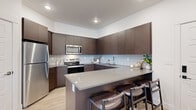 This thoughtfully designed 2-bedroom, 1-bathroom residence on a single floor delivers a cohesive blend of modern aesthetics and functional living. The open-concept layout seamlessly connects the kitchen, dining, and living areas, creating an inviting and efficient use of space throughout.

The kitchen is a standout feature, showcasing rich dark-brown cabinetry extending to the ceiling, a classic tile backsplash, stainless steel appliances including a refrigerator, range, and over-the-range microwave, and a generous quartz-topped peninsula with bar seating — ideal for casual dining or entertaining. Light wood-style flooring runs throughout, tying the spaces together with warmth and continuity.

The dining area sits adjacent to the kitchen and opens into the living room, which benefits from large windows that invite natural light and provide outdoor views. The hallway connects the common areas to the private quarters with clean, white-paneled doors and crisp trim work.

The primary bedroom offers a comfortable footprint, while the bathroom features a clean white vanity with ample drawer storage, a large mirror, and direct access to a spacious walk-in closet outfitted with multiple hanging rods and shelving. The overall ambiance is modern, bright, and move-in ready.