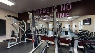 Located at 115 Carriage Hill Dr in Athens, Ohio, this single-story fitness center offers a comprehensive workout environment designed to accommodate a wide range of training needs. The space is organized into distinct functional zones, each outfitted with purpose-specific equipment and thoughtful design elements.

The strength training area is anchored by a multi-station cable machine, a Smith machine with a dedicated bench press setup, and a selection of selectorized machines targeting major muscle groups such as the legs and upper body. A tiered dumbbell rack lines one wall, offering a full range of free weights alongside battle ropes for functional training. Large mirrors span the strength zone, visually expanding the space and supporting proper form during exercises. A bold motivational phrase — "There Is No Finish Line" — is displayed prominently on a dark accent wall, setting an energetic tone throughout.

The cardio section features treadmills, a StairMaster step mill, and an elliptical trainer, positioned near windows that allow natural light and outdoor views of the surrounding landscape. Wall-mounted flat-screen televisions are positioned throughout the facility for entertainment during workouts.

Durable rubber flooring covers the entire space, and flush panel lighting provides consistent, bright illumination. Decorative artwork accents the neutral-toned walls, contributing to an engaging and motivating atmosphere.