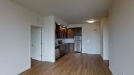 This single-floor space showcases a thoughtfully designed open-concept layout that seamlessly connects the kitchen and living areas. The kitchen is outfitted with dark espresso-toned cabinetry, offering both upper and lower storage, complemented by open shelving for display or additional accessibility. Stainless steel appliances — including a refrigerator, microwave, and dishwasher — provide a sleek, contemporary aesthetic. A built-in sink with recessed lighting above the kitchen area adds functional illumination to the workspace.

The flooring throughout is a warm, medium-toned hardwood-style material that adds continuity and a sense of warmth across the open space. White walls and a neutral ceiling palette keep the interior feeling clean and bright, enhanced by flush-mounted ceiling lighting.

The entryway features a solid front door with a modern hardware finish, providing a clean and secure point of entry. An additional interior door, visible to the left, offers access to a separate room or closet, adding privacy and storage versatility. Overall, the space presents a clean, contemporary design with functional finishes suited for everyday living.