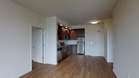 This single-floor space showcases a thoughtfully designed open-concept layout that seamlessly connects the kitchen and living areas. The kitchen is outfitted with dark espresso-toned cabinetry, offering both upper and lower storage, complemented by open shelving for display or additional accessibility. Stainless steel appliances — including a refrigerator, microwave, and dishwasher — provide a sleek, contemporary aesthetic. A built-in sink with recessed lighting above the kitchen area adds functional illumination to the workspace.

The flooring throughout is a warm, medium-toned hardwood-style material that adds continuity and a sense of warmth across the open space. White walls and a neutral ceiling palette keep the interior feeling clean and bright, enhanced by flush-mounted ceiling lighting.

The entryway features a solid front door with a modern hardware finish, providing a clean and secure point of entry. An additional interior door, visible to the left, offers access to a separate room or closet, adding privacy and storage versatility. Overall, the space presents a clean, contemporary design with functional finishes suited for everyday living.