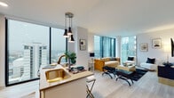 This single-floor urban residence is defined by its striking floor-to-ceiling windows, which frame expansive city skyline views from multiple rooms and flood the interior with natural light throughout the day. Light hardwood flooring runs continuously through the open-concept living spaces, reinforcing a cohesive, airy aesthetic.

The kitchen is thoughtfully designed with white shaker cabinetry, a subway tile backsplash, stainless steel appliances, and a central island with seating. A brass-finished sink and trio of pendant lights above the island add refined touches. The kitchen connects seamlessly to the living area, where corner windows provide panoramic urban vistas, making it an inviting space for both relaxing and entertaining.

Two bedrooms each benefit from large floor-to-ceiling windows overlooking the cityscape. One bedroom includes a walk-in closet with built-in shelving, paired with an en-suite bathroom featuring a glass-enclosed walk-in shower, hexagonal tile flooring, and a wood-toned vanity with an undermount sink. A well-proportioned hallway, adorned with curated artwork, connects the living spaces to the private quarters, enhancing the overall sense of thoughtful design throughout.