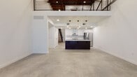 This two-story industrial-modern loft masterfully blends raw architectural character with contemporary finishes. The main level showcases expansive polished concrete floors throughout the open-plan living and kitchen areas, with soaring ceilings that reveal exposed wooden beams and structural elements overhead. A sleek kitchen serves as the centerpiece, featuring a bold dark-toned island with white quartz countertops, a subway tile backsplash, stainless steel appliances including a gas range with a wall-mounted hood, and pendant lighting that adds warmth to the space. An open metal railing staircase connects the two floors, offering clear sightlines across the entire main level. The upper mezzanine level is finished with plush carpeting and is defined by dramatic exposed wood beam ceilings and eye-catching dark steel structural trusses that speak to the building's industrial heritage. A sliding barn door on the upper level adds a functional yet stylish touch. Large grid-paned windows on the main level fill the space with natural light. The bathroom is cleanly appointed with white subway tile, a tub-shower combination, marble-look tile flooring, and matte black hardware accents, completing the space's cohesive industrial-modern aesthetic.
