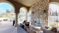 Located at 3875 Main in Chula Vista, this outdoor clubhouse offers a well-appointed communal gathering space designed for both relaxation and entertaining. The covered lounge area is anchored by a striking floor-to-ceiling river rock fireplace, framed by wrought iron arched gates that allow natural light and garden views to flow through. Comfortable wicker seating with plush cushions in neutral tones and green accents surrounds a wooden coffee table, creating an inviting social atmosphere. Elegant stucco arches supported by cylindrical columns define the perimeter of the covered area, seamlessly connecting the sheltered lounge to the open-air patio beyond. The exterior patio is generously sized and features multiple dining tables with patio chairs shaded by large umbrellas, providing ample space for group gatherings. A BBQ grill station and fire pit add functional outdoor cooking and lounging options. The surrounding landscaping includes layered, drought-tolerant plantings and stone retaining walls that lend a natural, polished character to the space. Aggregate concrete flooring and warm-toned stucco walls carry a cohesive Mediterranean-inspired aesthetic throughout. Wall-mounted lantern-style light fixtures add a refined finishing touch to this well-designed community outdoor retreat.
