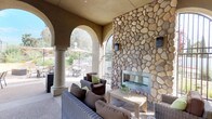 Located at 3875 Main in Chula Vista, this outdoor clubhouse offers a well-appointed communal gathering space designed for both relaxation and entertaining. The covered lounge area is anchored by a striking floor-to-ceiling river rock fireplace, framed by wrought iron arched gates that allow natural light and garden views to flow through. Comfortable wicker seating with plush cushions in neutral tones and green accents surrounds a wooden coffee table, creating an inviting social atmosphere. Elegant stucco arches supported by cylindrical columns define the perimeter of the covered area, seamlessly connecting the sheltered lounge to the open-air patio beyond. The exterior patio is generously sized and features multiple dining tables with patio chairs shaded by large umbrellas, providing ample space for group gatherings. A BBQ grill station and fire pit add functional outdoor cooking and lounging options. The surrounding landscaping includes layered, drought-tolerant plantings and stone retaining walls that lend a natural, polished character to the space. Aggregate concrete flooring and warm-toned stucco walls carry a cohesive Mediterranean-inspired aesthetic throughout. Wall-mounted lantern-style light fixtures add a refined finishing touch to this well-designed community outdoor retreat.