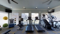 This single-story fitness center in Flower Mound is designed to support an active lifestyle with a focused, functional layout. The space is outfitted with a selection of cardio machines, including treadmills, an upright stationary bike, and a stair climber, all arranged to maximize usability and flow. A set of medicine balls mounted along the wall adds a strength and conditioning element to the room. The interior is finished with neutral gray walls and recessed lighting, creating a clean and practical atmosphere. Multiple large windows fitted with horizontal blinds line the walls, allowing ample natural light to filter into the space while offering views of the surrounding neighborhood. The patterned carpet flooring adds a subtle design element underfoot. Ceiling-mounted ventilation ensures comfortable air circulation throughout workouts. Overall, the fitness center offers a well-lit, organized environment with essential equipment for cardiovascular training and light functional fitness routines.