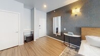 Located at 5501 E 18th St in Sioux Falls, South Dakota, this thoughtfully designed single-floor suite blends contemporary aesthetics with practical functionality. The space is unified by warm luxury vinyl plank flooring and a carefully curated palette of soft neutrals accented by bold, textured wallcoverings.

The bedroom serves as the centerpiece of the suite, featuring a distinctive marbled topographic-patterned accent wall adorned with elegant gold sconce lighting. A large sliding glass door floods the room with natural light while providing direct outdoor access. A wall-mounted flat-screen television, a cozy sitting area, and ample space complete the comfortable retreat. A vintage-style area rug adds warmth underfoot.

The entryway makes a strong first impression with a dramatic dark topographic-patterned wallcovering, a full-length wall mirror, a console table, and a compact refrigerator — offering both style and convenience upon arrival.

The private bathroom is designed with accessibility in mind, equipped with sturdy grab bars near the toilet and shower area. A rich golden geometric-patterned accent wall brings visual interest, while a dark wood vanity topped with a marble-look surface adds a refined touch. Framed artwork and thoughtful details round out the space.

Overall, this suite offers a polished, hotel-inspired atmosphere with modern design elements and accessible features.