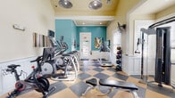 This fitness center offers a thoughtfully designed workout environment that balances both style and functionality. The space is defined by its elevated ceilings, which lend an open, airy feel, and are complemented by large industrial-style pendant lights that provide ample illumination alongside recessed ceiling fixtures. A striking teal accent wall anchors the cardio area, adding a vibrant pop of color that energizes the space, while creamy yellow upper walls and classic white wainscoting create a polished, cohesive aesthetic throughout.

The flooring features a bold tan and charcoal checkered pattern that adds a dynamic visual element underfoot. The cardio zone is well-stocked with a row of elliptical trainers and treadmills, including Matrix-brand equipment, each outfitted with individual monitors. Spin bikes are also available for cycling workouts. A dedicated strength training area includes a multi-functional cable machine, a dumbbell rack with a full range of free weights, a weight bench, and wall-mounted televisions for entertainment during workouts.

Natural light filters in through multiple windows dressed with white blinds, offering views of the surrounding greenery. A "Cardio Theater Rules" sign near the entrance sets the tone for a community-minded environment. The space connects to an adjacent corridor, suggesting it is part of a larger amenity facility.