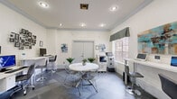 Located at 5009 Park Central Dr in Orlando, FL, this single-floor community business center offers a thoughtfully designed space for work and collaboration. The layout flows seamlessly from an elegant reception lounge into a functional business suite. The lounge area sets a refined tone with a tray ceiling, a statement chandelier, large floor planters with tropical greenery, plush white armchairs, a colorful area rug, and glass-paneled French doors that provide a visual connection to the work area beyond. The business suite is finished with slate-tiled flooring in a classic checkerboard pattern and is lined with white walls accented by crown molding. Multiple computer workstations with adjustable bar-height seating are arranged along the perimeter, complemented by a multifunction printer and copier for full office capability. A round white table with modern wire chairs serves as a central gathering point, suitable for informal meetings or group work. Large windows dressed with patterned valances allow natural light to fill the room while offering views of the surrounding landscape. Decorative wall art and potted plants add warmth to the otherwise crisp, professional aesthetic. Recessed lighting throughout ensures consistent illumination across the entire space.