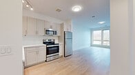 This contemporary two-floor residence showcases a clean, modern aesthetic throughout, with light hardwood flooring and crisp white walls creating a bright, airy ambiance. The well-appointed kitchen features shaker-style cabinetry in a soft grey-white finish, a textured tile backsplash, white countertops, and a full suite of stainless steel appliances including a range, microwave, dishwasher, and refrigerator. Track lighting and an open layout connect the kitchen seamlessly to the living area, where large windows allow generous natural light to pour in. The staircase, finished with a sleek black metal railing and light wood treads, connects the two floors with a contemporary touch visible in the dollhouse view. Upper-level living areas are generously sized with tall ceilings and multiple large windows framing views of the surrounding neighborhood. One room opens directly onto a private outdoor balcony through sliding glass doors, offering outdoor space with scenic rooftop and treetop views. Bathrooms are finished with large-format tile flooring; one features a glass-enclosed walk-in shower with marble-style tile surround, a vanity with stone countertop, and brushed nickel fixtures, while another is a clean, accessible half-bath. Throughout, the space exudes a fresh, modern character suited for comfortable everyday living.