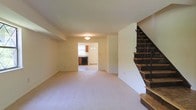 This two-story residence offers a practical and comfortable layout spread across two well-connected floors. The main level greets visitors with a generously sized open living area finished with plush wall-to-wall carpeting that lends a warm, inviting feel to the space. Neutral-toned walls and a flush ceiling light fixture keep the area bright and versatile, ready to accommodate a variety of furnishing arrangements.

A staircase with classic wooden treads and a distinctive wrought iron railing runs along one side of the main floor, providing both a functional and decorative focal point as it leads to the upper level. The open-plan flow guides naturally toward the kitchen area, which is visible through a connecting passage. The kitchen features warm wood-toned cabinetry, a dark appliance suite, and the benefit of natural light from a window — offering a functional workspace with a welcoming atmosphere.

The entry foyer is positioned adjacent to the kitchen, framed by a paneled front door that allows additional natural light to filter into the home. Throughout the main floor, the seamless transition between spaces creates an easy, open circulation that makes the home feel cohesive and well-proportioned. The second floor, accessible via the staircase, expands the home's living capacity further, offering additional private spaces above the main living level.