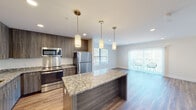 This thoughtfully designed single-floor unit offers a seamless blend of modern finishes and functional living spaces. At the heart of the home is an open-concept kitchen that flows directly into the living area, creating an inviting and connected layout. The kitchen is appointed with dark wood-toned cabinetry, a full suite of stainless steel appliances including a refrigerator, range, microwave, and dishwasher, and a striking granite-topped island with pendant lighting above. A trio of cylindrical pendant lights adds a warm, contemporary touch to the space.

The living area is bright and generously proportioned, featuring recessed lighting, large windows dressed with vertical blinds, and a sliding glass door that opens to an outdoor seating area. Hardwood-style flooring runs consistently throughout the unit, lending a cohesive and polished look.

The bedrooms are quiet and well-lit, each benefiting from large windows that invite natural light and offer views of the surrounding greenery. Clean white walls and neutral tones create a calm, versatile backdrop throughout.

The bathroom continues the unit's design language with a granite-topped vanity, dark wood cabinetry, and wood-style flooring, maintaining a consistent aesthetic that ties the entire space together elegantly.