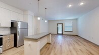 This well-appointed one-bedroom, one-bathroom unit in Leander, TX offers a thoughtfully designed single-level layout that maximizes functionality and comfort. The open-concept kitchen and living area serves as the heart of the home, anchored by a granite-topped island with elegant pendant lighting overhead. The kitchen is outfitted with crisp white upper cabinetry, warm wood-toned lower cabinets, a classic subway tile backsplash, and a full suite of stainless steel appliances including a refrigerator, range, microwave, and dishwasher. Light wood-style flooring flows throughout the main living areas, lending a bright and airy feel. The living room benefits from natural light through a glass-paneled door and an adjacent window fitted with blinds. Built-in open shelving adds a practical and stylish accent near the entryway. The bedroom features plush wall-to-wall carpet, a ceiling fan, and dual windows that invite natural light. Two closets provide generous storage, and the bathroom is finished with a wood-toned vanity, granite countertop, tile flooring, and a tiled tub-shower combination. A dedicated laundry area completes this efficient and modern living space.