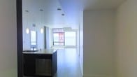 This single-floor space showcases a sleek, contemporary design characterized by an open-plan layout that seamlessly connects the kitchen and living areas. The kitchen is equipped with a dark-toned island unit, stainless steel dishwasher, and a light-colored countertop, complemented by a stylish pendant light that adds a focused decorative element overhead. The flooring throughout is a light-toned hardwood, contributing to the clean, minimalist feel of the interior. Large floor-to-ceiling windows at the far end of the living area flood the space with abundant natural light while framing views of the surrounding urban environment. The white walls and white ceiling further enhance the brightness and sense of openness throughout. Recessed and flush-mount ceiling lighting supplements the natural light, ensuring the space remains well-lit at all times. A corridor extends toward a private area of the unit, suggesting additional rooms beyond the main living space. Overall, the space conveys a polished, modern ambiance with a neutral palette and well-considered architectural details.